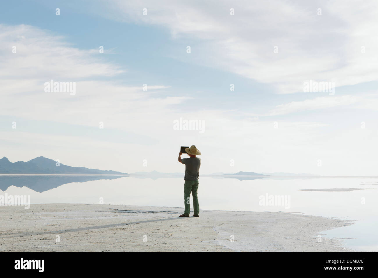 Un uomo in piedi sul bordo dell'allagato Bonneville Saline al tramonto, scattare una fotografia con un dispositivo tablet pc. Foto Stock