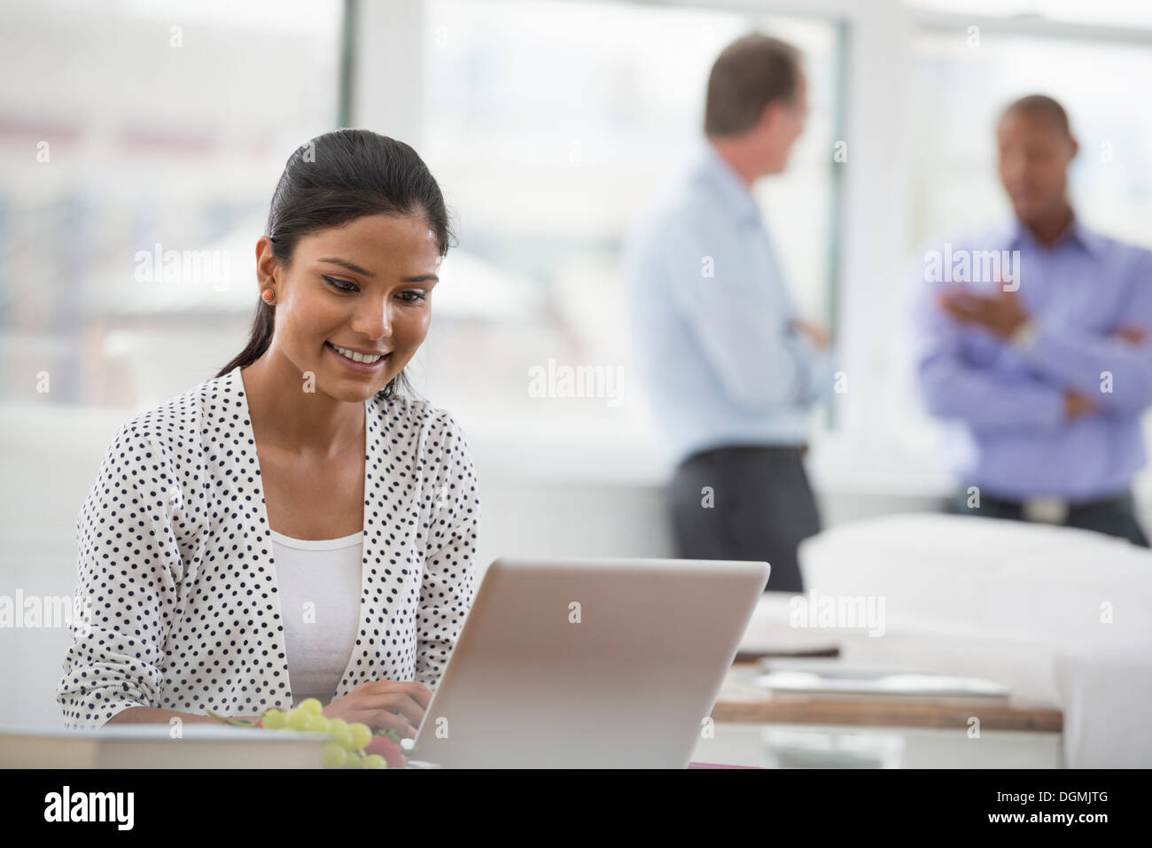 Vita in ufficio. Una donna seduta a una scrivania utilizzando un computer portatile. Due uomini in background. Foto Stock