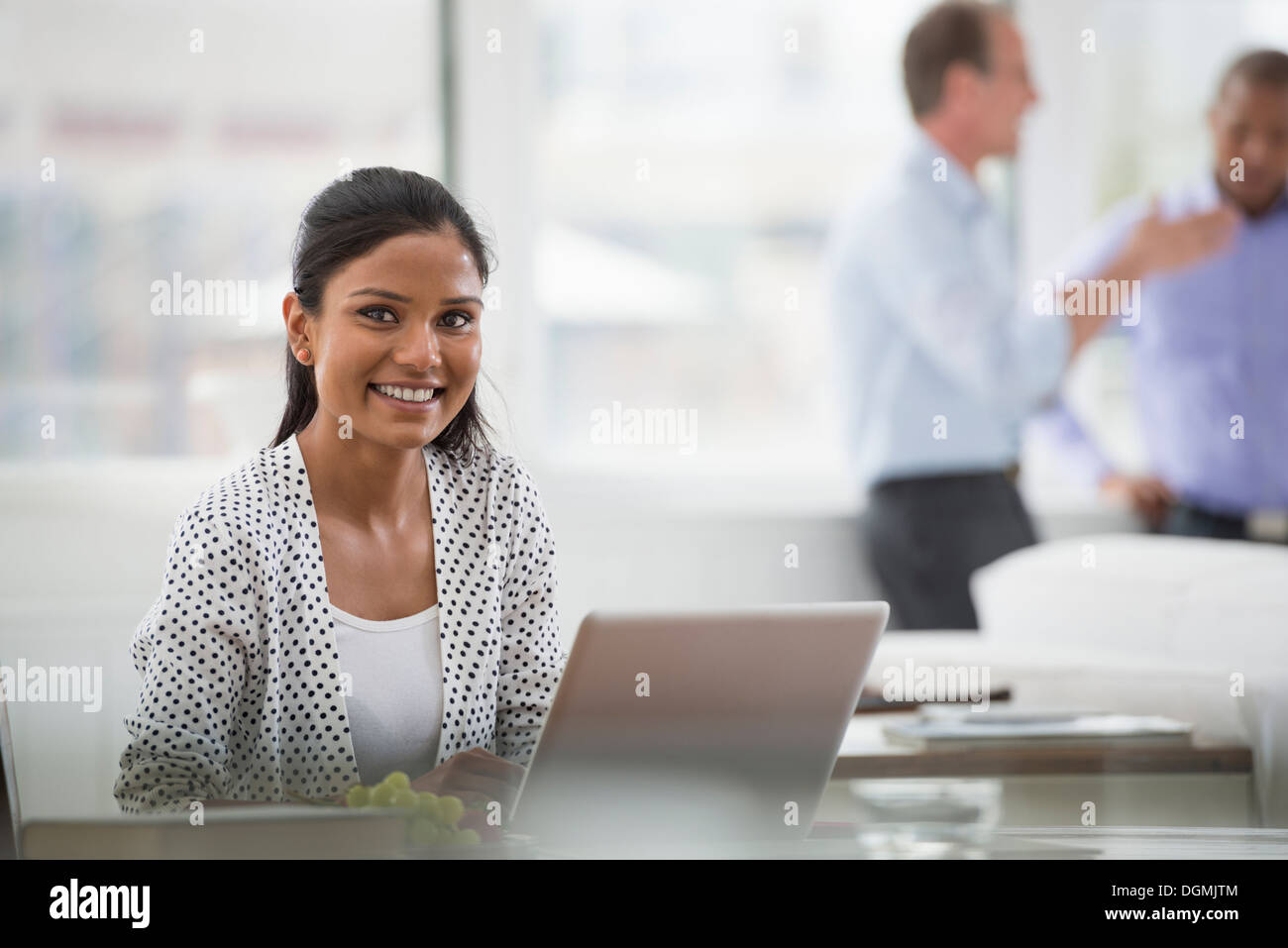 Vita in ufficio. Una donna seduta a una scrivania utilizzando un computer portatile. Due uomini in background. Foto Stock