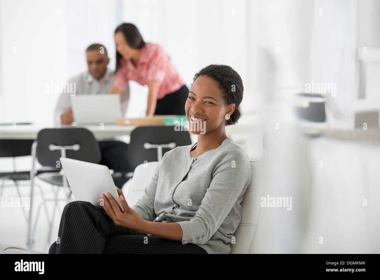 Business. Una donna seduta utilizzando una tavoletta digitale. Due persone in background guardando lo schermo di un computer. Foto Stock