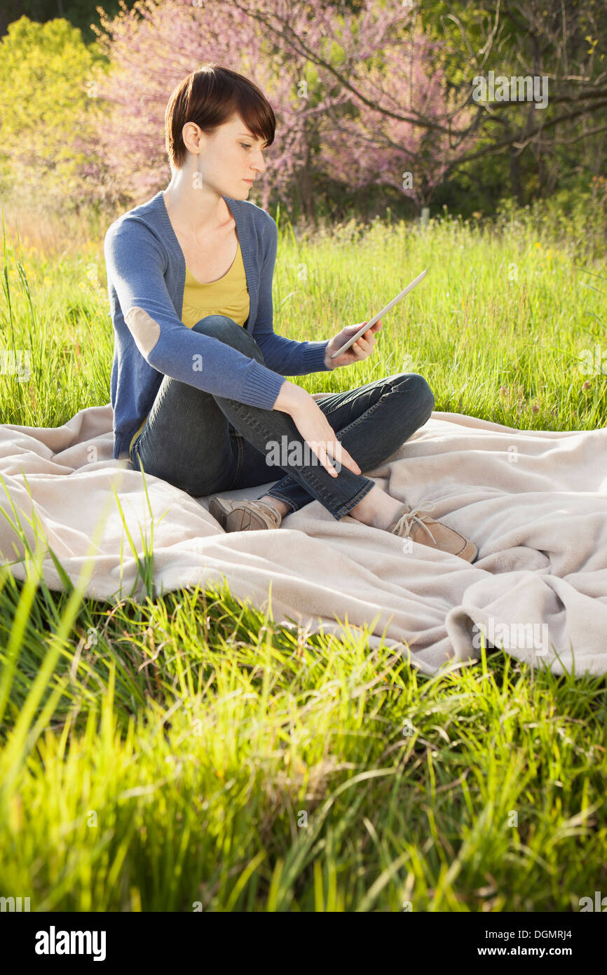 Una giovane donna seduta in un campo su una coperta, detenzione e guardando lo schermo di una tavoletta digitale. Lavorare all'esterno. Foto Stock