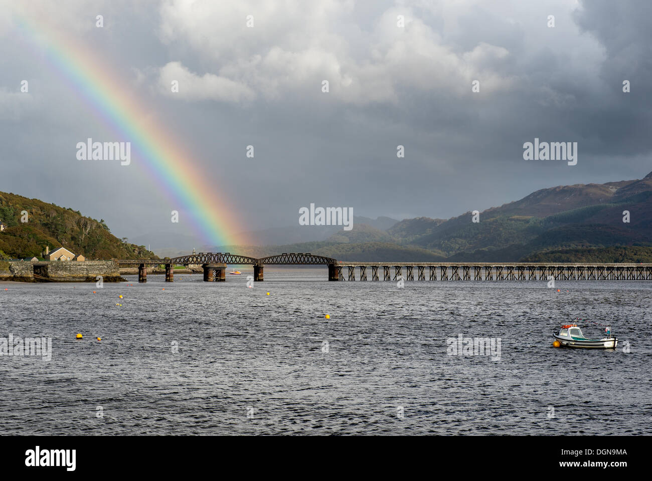 La bocca del Mawddach estuary in Galles del nord è attraversata dalla Barmouth Bridge. Un arcobaleno termina appena sotto uno degli archi oscillante accensione RNLI una scialuppa di salvataggio. Aperto nel 1867 è costruito in legno e ferro. Parte del ponte è stato ricostruito nel 1901 a swing round per consentire il passaggio di Tall Ships. Fu costruito per portare una sola via la stazione ferroviaria e la linea Cambriano lo utilizza per questo giorno. A fianco della stazione ferroviaria si trova un sentiero per pedoni. Foto Stock