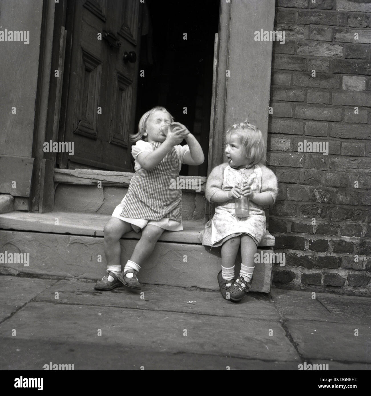 Immagine storica,anni cinquanta di due bambine seduto sulla porta di casa urbana, uno di bere dalla sua bottiglia, gli altri a guardare. Foto Stock