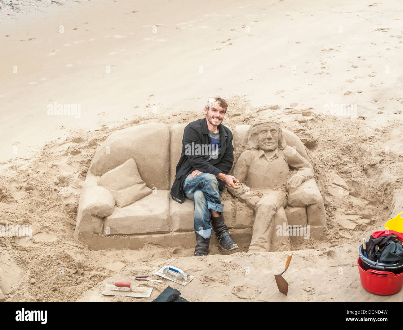 Scultore sorridente in posa, creando la scultura di sabbia di un uomo disteso su un divano, sul Fiume Tamigi South Bank di Londra, UK per suggerimenti Foto Stock