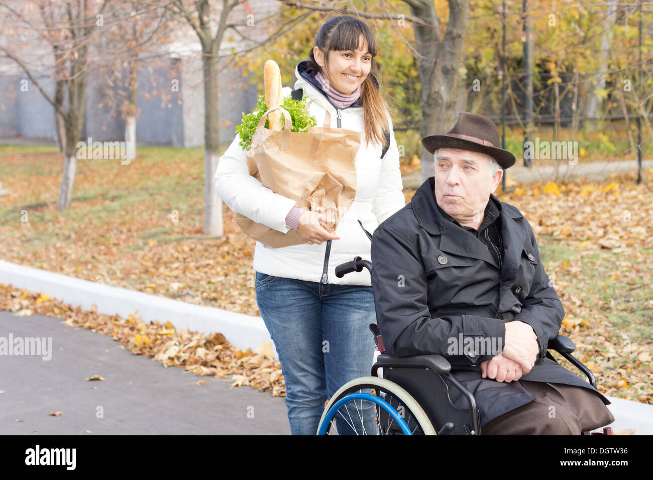 Donna aiutare un anziano uomo disabili con la sua spesa spingendo la sua sedia a rotelle lungo la strada e portando la sua borsa di Foto Stock