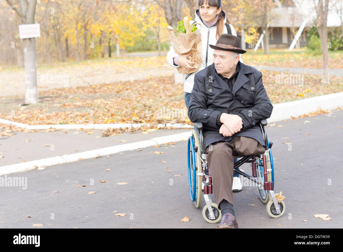 Senior uomo disabili di essere aiutato con il suo shopping seduto nella sua sedia a rotelle come un caregiver femmina o la figlia lo spinge lungo il Foto Stock