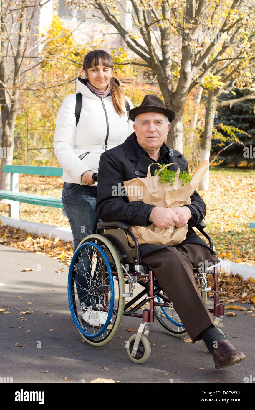 Senior uomo disabile andando a fare la spesa ha aiutato da un sorridente donna attraente che sta spingendo la sua sedia a rotelle lungo la strada come egli detiene il pacchetto di generi alimentari. Foto Stock