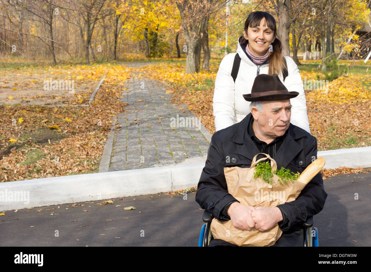 Donna sorridente spingendo un vecchio su una sedia a rotelle di ritorno da una passeggiata attraverso un colorato parco autunno con una grande borsa di Foto Stock