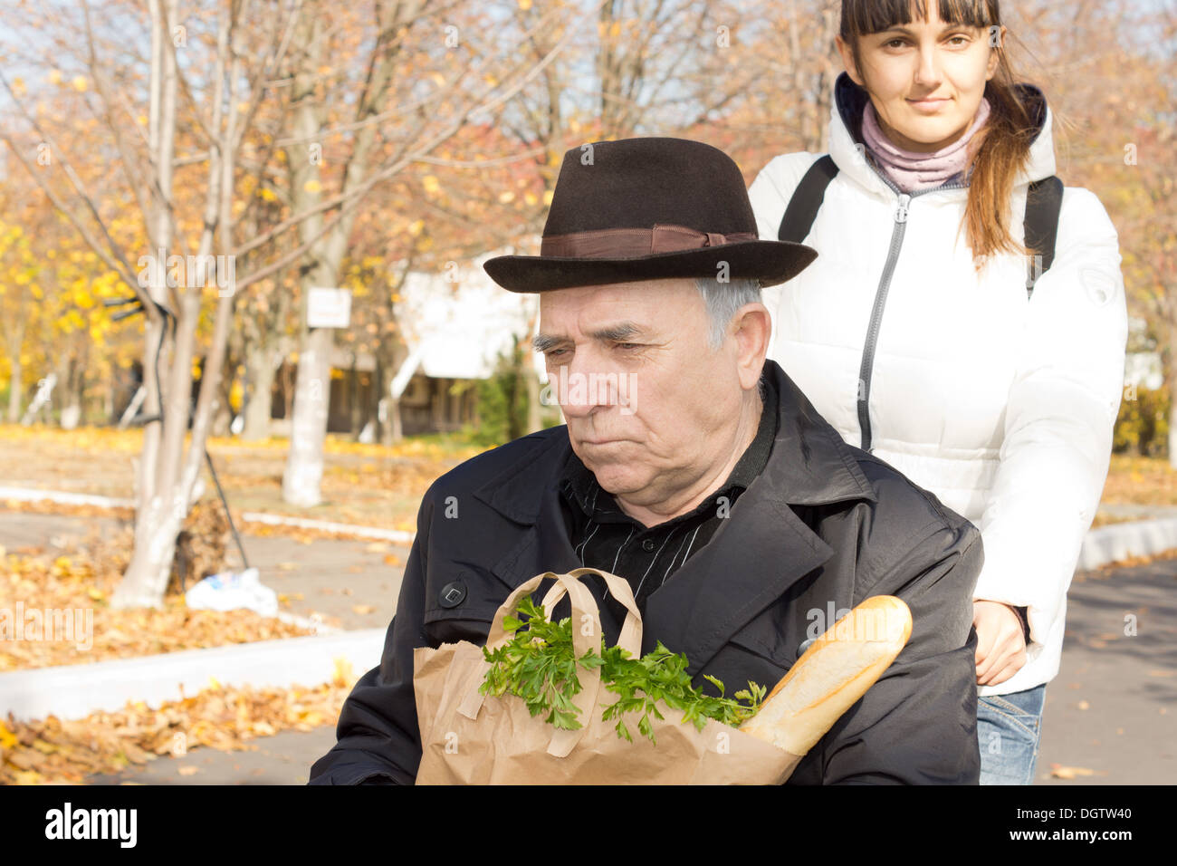 Attraente di mezza età caregiver spingendo un anziano uomo disabili nella sua sedia a rotelle come egli detiene un pacchetto di generi alimentari freschi sul suo giro dopo andare a fare shopping. Foto Stock