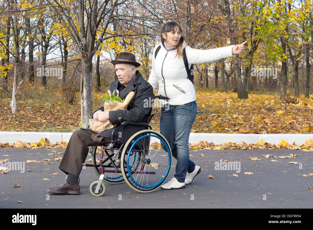 Donna spingendo un uomo anziano in una sedia a rotelle e stirando il suo braccio rivolto a qualcosa e a chiamare in quanto a piedi lungo la strada. Foto Stock