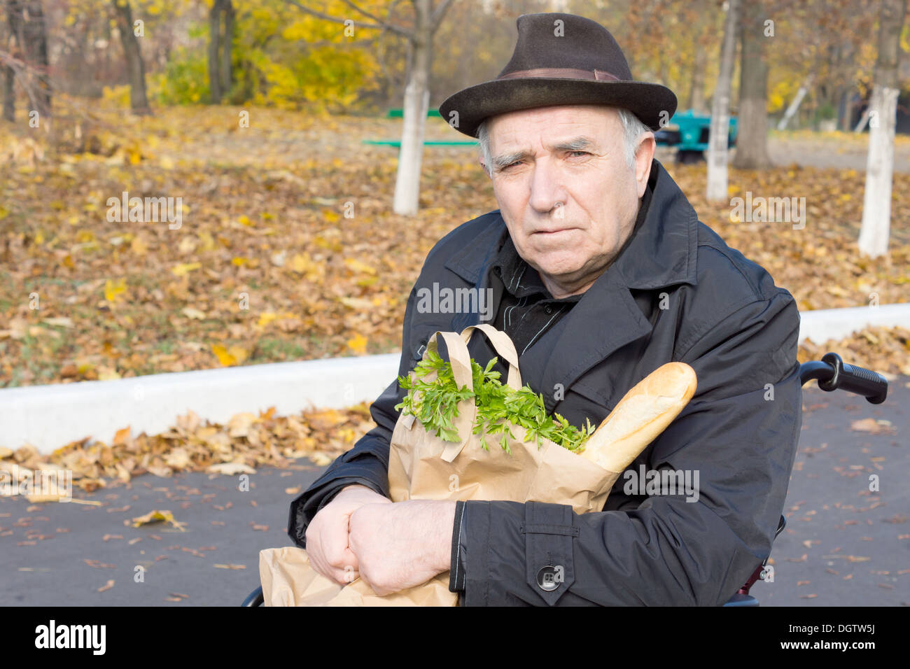 Senior uomo seduto in una sedia a rotelle con un sacchetto di carta di generi alimentari innestata sul suo giro come egli attende in strada per assistenza. Foto Stock