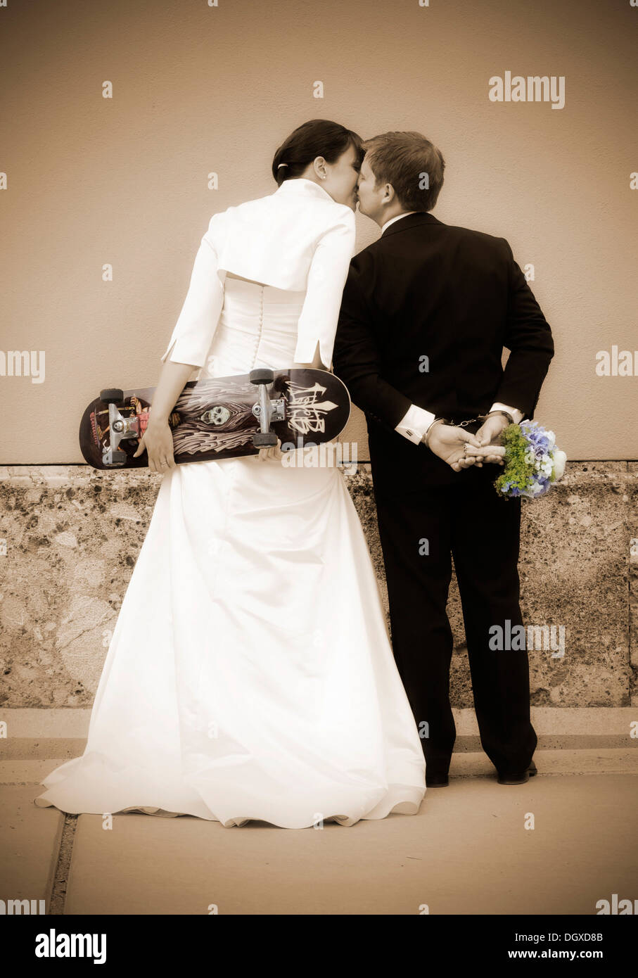 Kissing sposi visto da dietro la sposa è in possesso di uno skateboard, lo sposo indossa le manette e tenendo un bridal Foto Stock