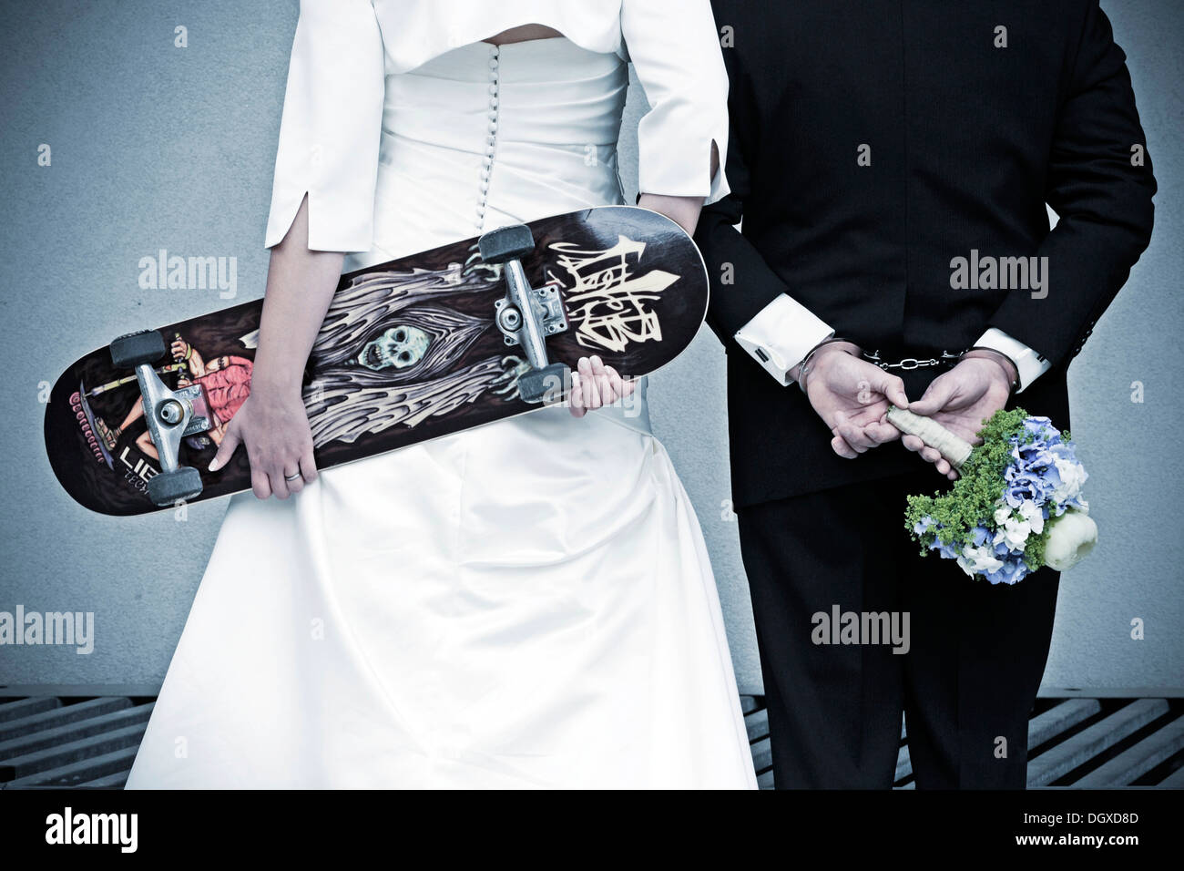 Coppie in viaggio di nozze visto da dietro la sposa è in possesso di uno skateboard, lo sposo indossa le manette e tenendo un bouquet nuziale Foto Stock