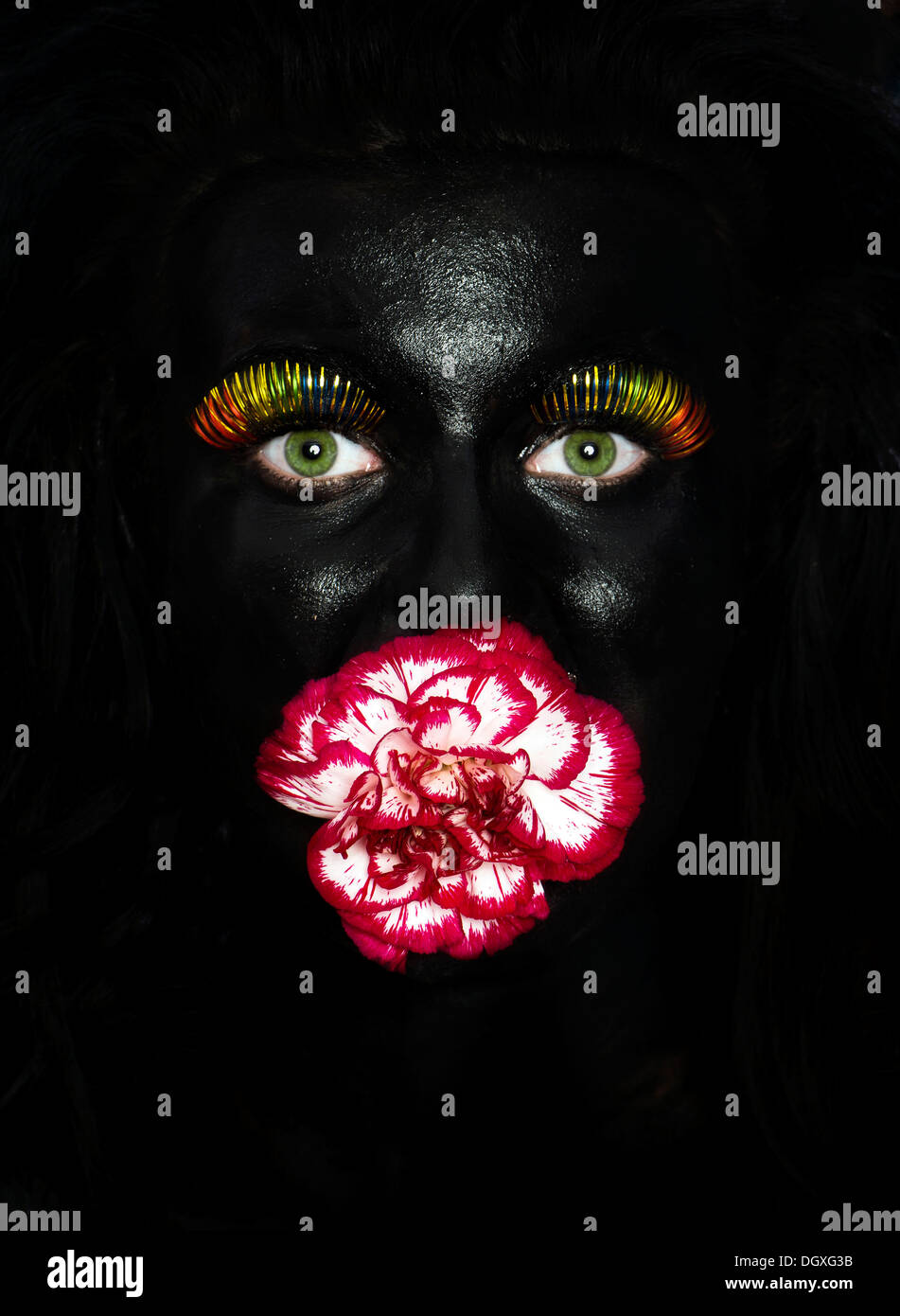 Volto di una giovane donna dipinta di nero facepaint, con multi-colore di ciglia e un fiore nella sua bocca, Austria Foto Stock