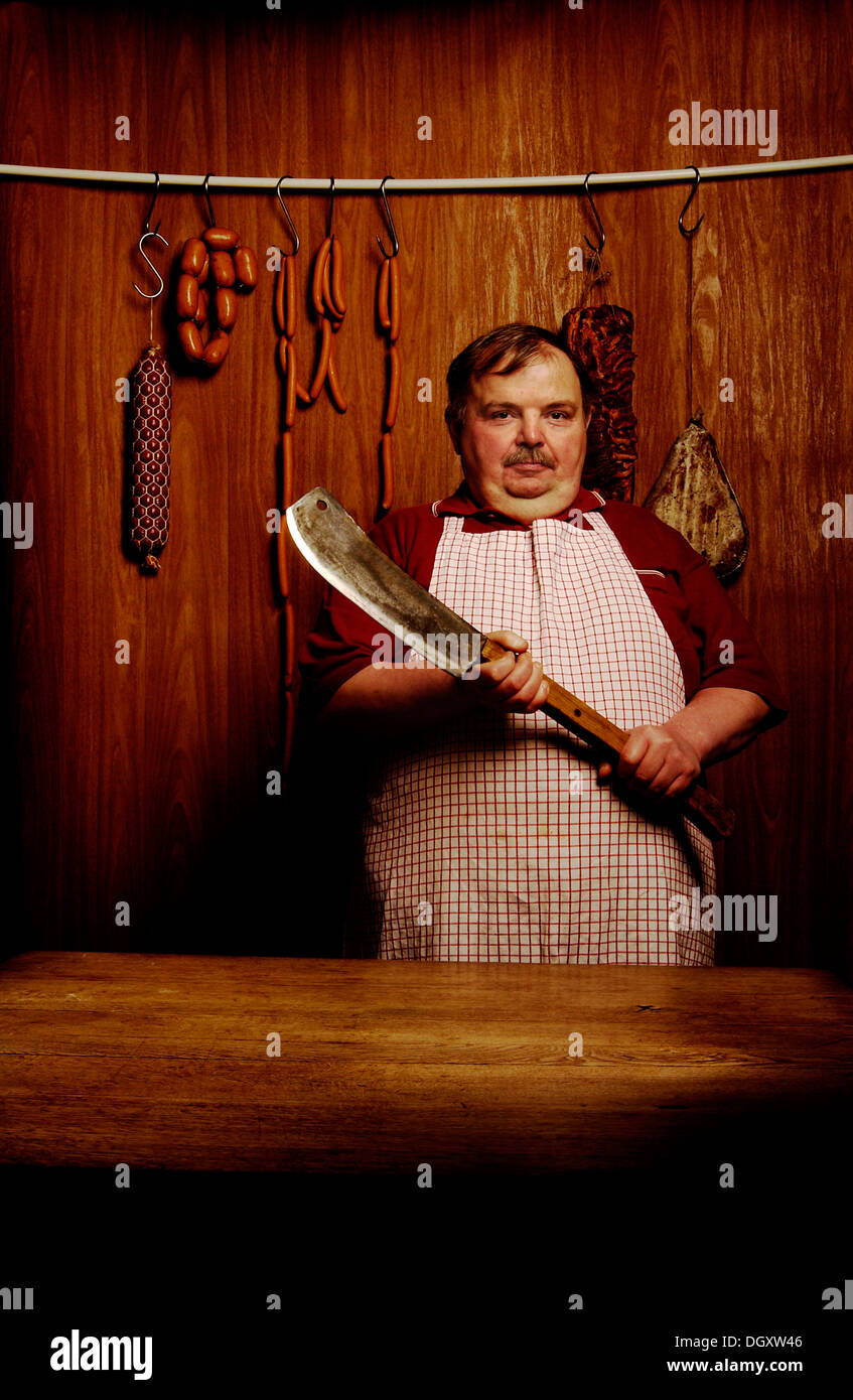 Fat butcher tenendo una mannaia di carne in una macelleria in negozio con un ambiente in legno Foto Stock