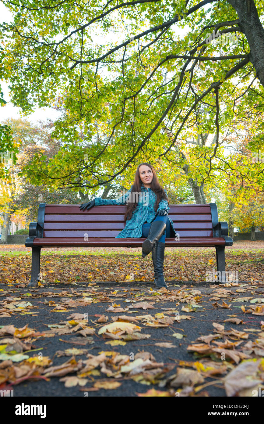 Giovane donna in un parco durante l'autunno seduta su una panchina con foglie sul terreno in primo piano e gli alberi in background Foto Stock