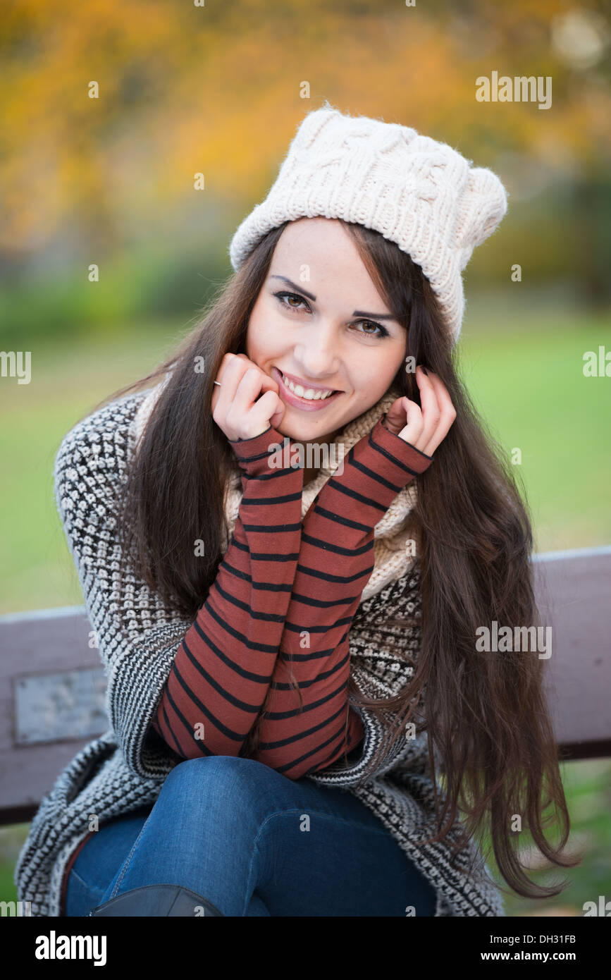 Giovane donna in un parco durante l'autunno seduta su una panchina indossando un cappello wooly sorridente verso la telecamera con fiducia Foto Stock