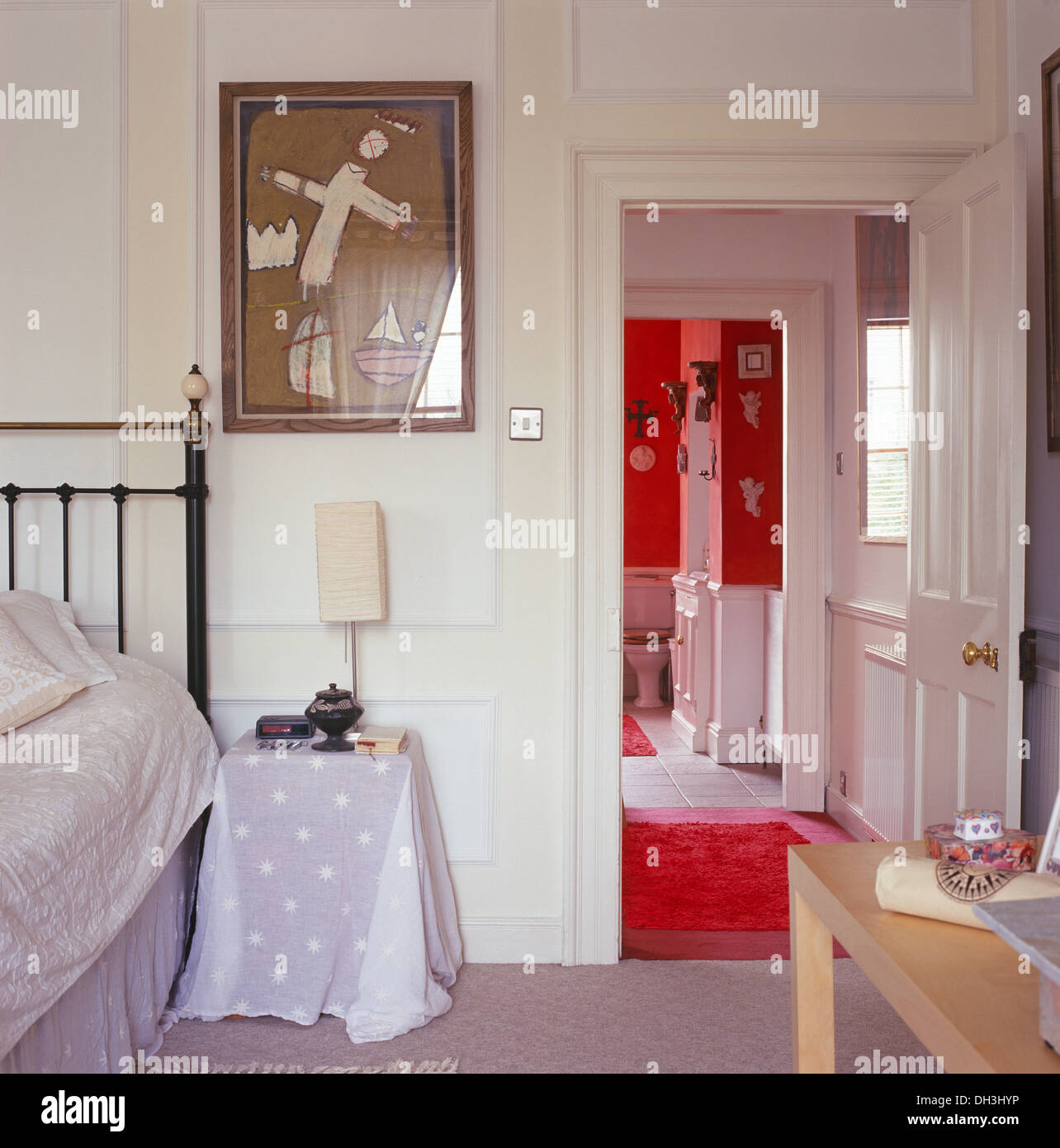 Bianco camera da letto con pannelli con foto sopra il comodino con panno bianco accanto a porta aperta al rosso bagno en-suite Foto Stock