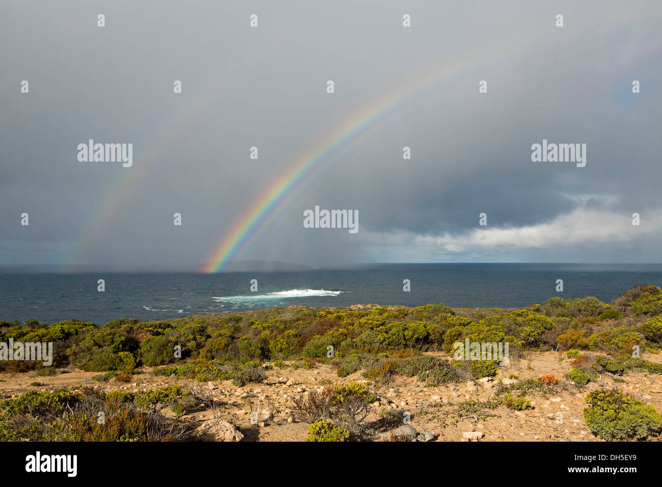 Ampia e vivacemente colorato rainbow affettatura attraverso il grigio e il cielo tempestoso e in oceano scuro con onde bianco Foto Stock