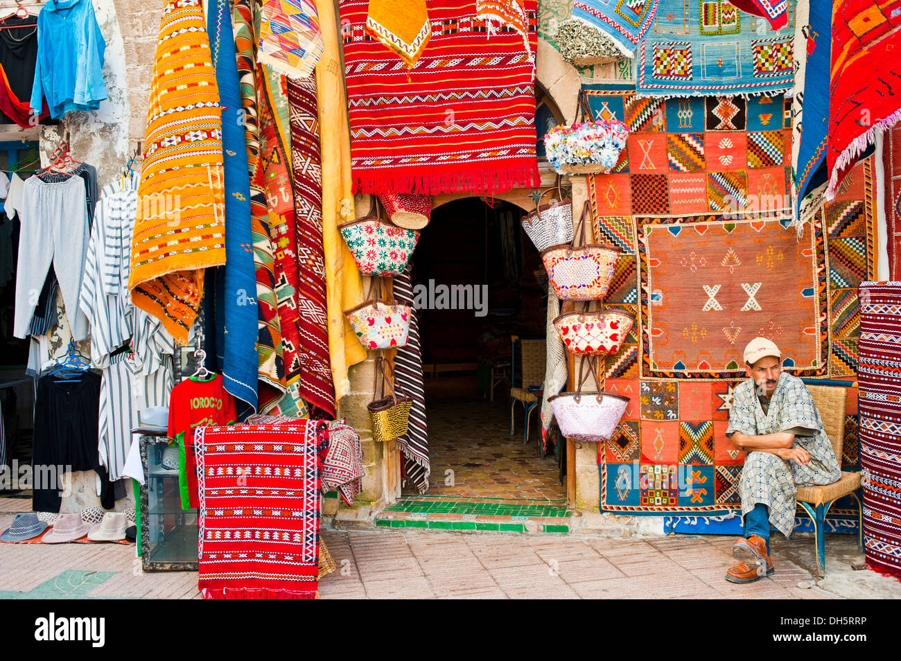 Un commerciante di tappeti seduto di fronte al suo negozio nel souk o bazaar, moquette o tappeti sono appesi alla parete per la vendita, Essaouira Foto Stock