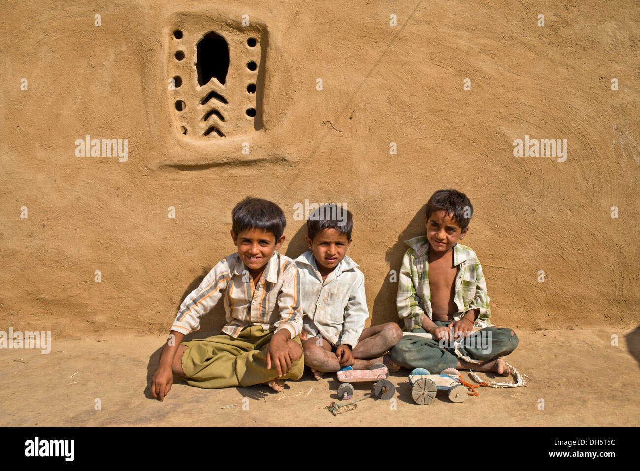 Tre ragazzi seduti al di fuori di una casa di fango, fatti in casa a base di modellini di auto di fronte a loro, Wüste Thar, Rajasthan, India Foto Stock