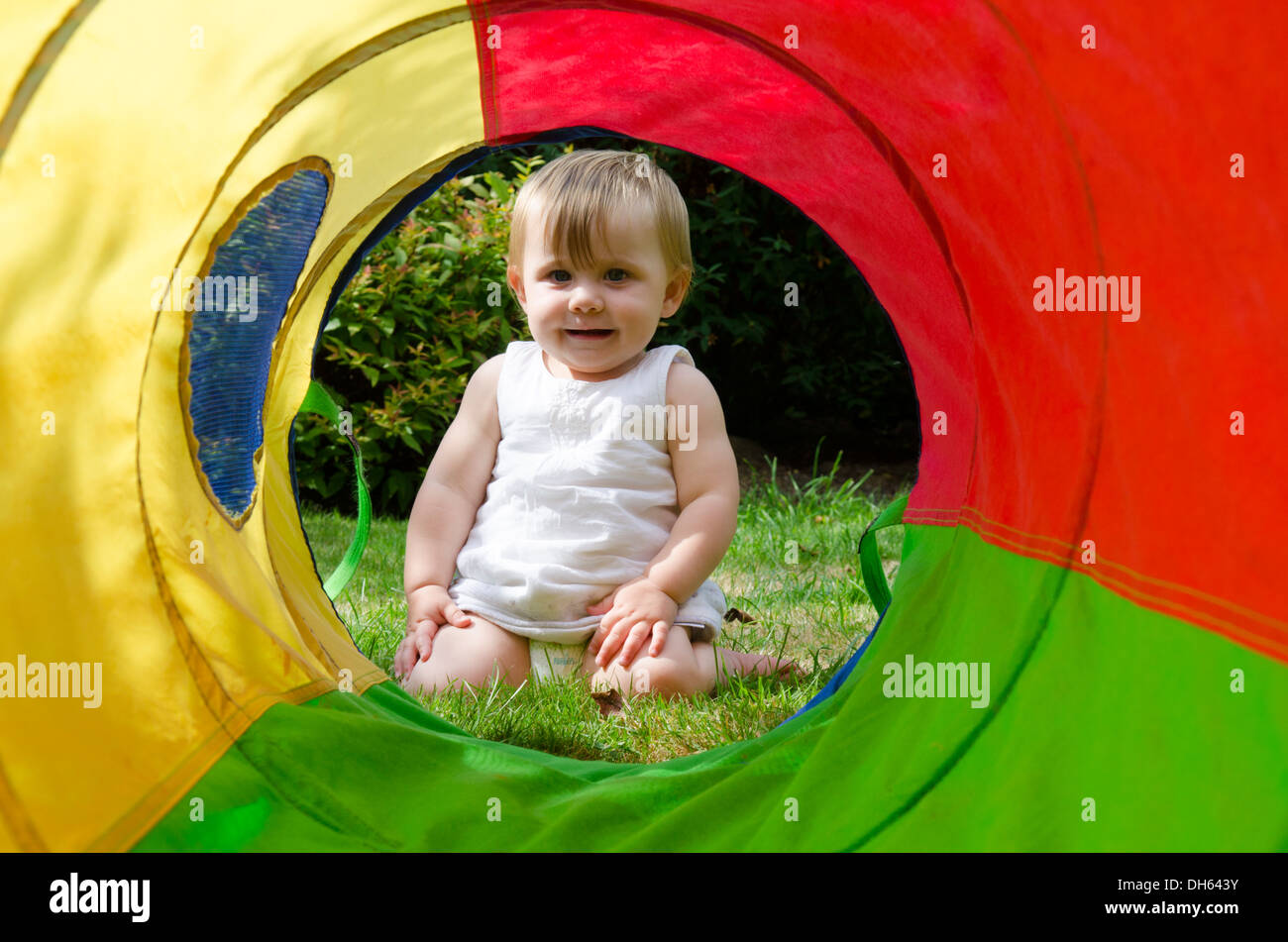 Nove mesi ragazza in giardino guardando attraverso il grande panno colorati Tubo giocattolo. Agosto. Regno Unito. Foto Stock