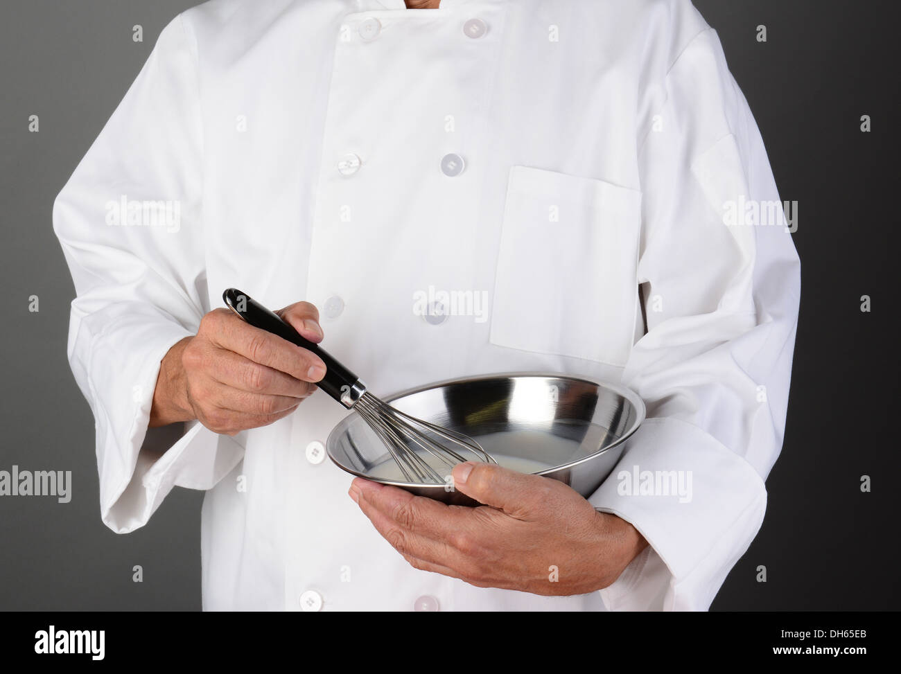 Primo piano di uno Chef tenendo un acciaio inossidabile recipiente di miscelazione e una frusta. Formato orizzontale su una luce di sfondo grigio scuro. Foto Stock