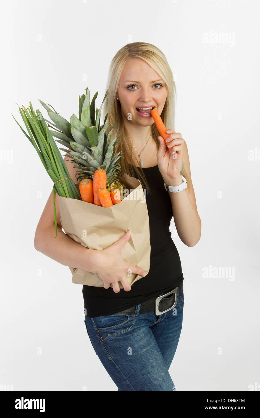 Giovane donna con un sacchetto pieno di frutta e verdura nel suo braccio, tenendo una carota in mano Foto Stock