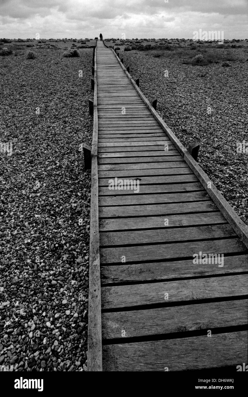 In bianco e nero di percorso di legno sulla spiaggia ghiaiosa di scomparire verso l'orizzonte con persona in distanza a Dungeness Foto Stock