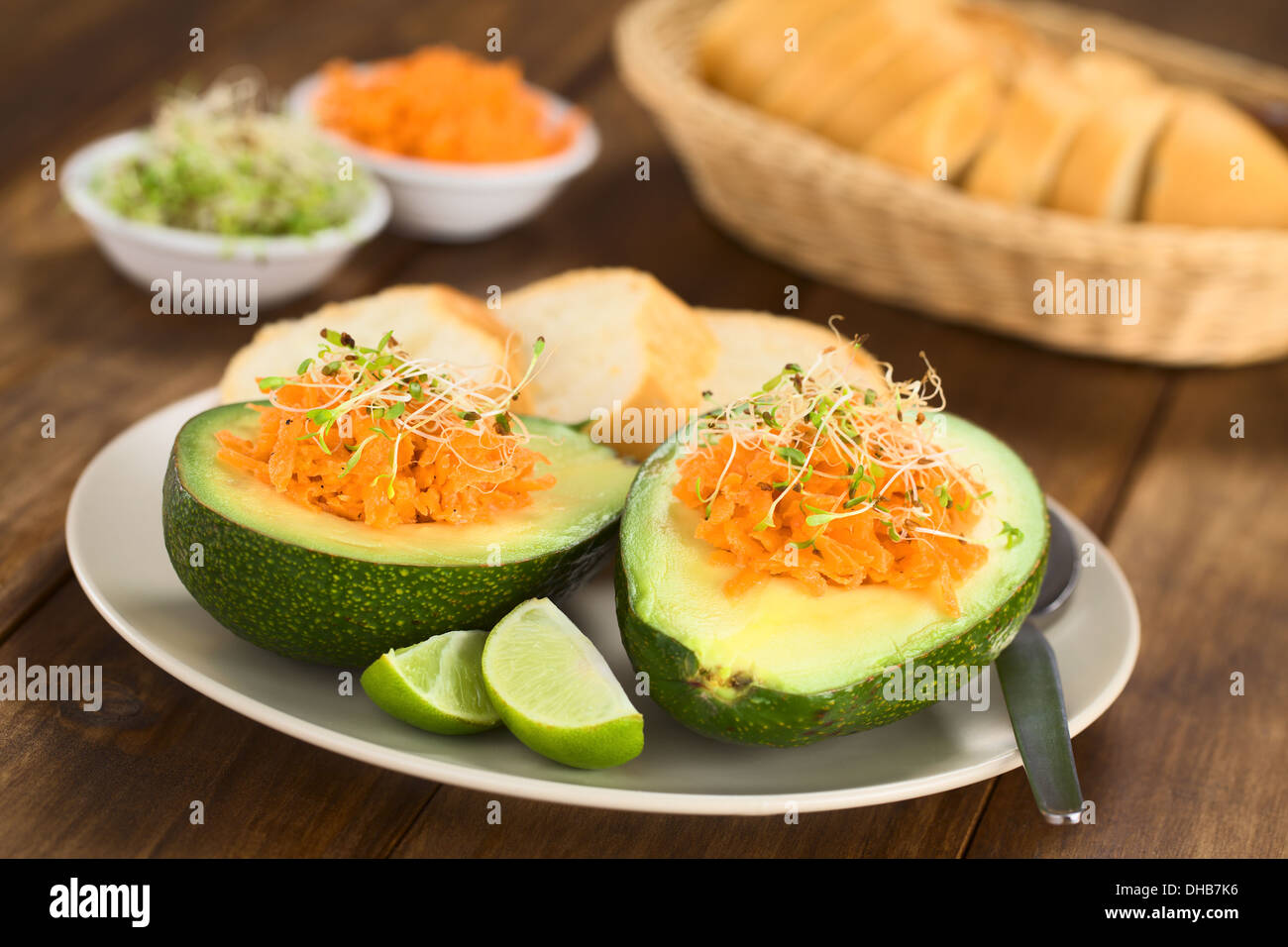 Metà di Avocado riempito con carote grattugiate e cosparso di germogli alfalfa servita su piastra con spicchi di lime e baguette fette Foto Stock