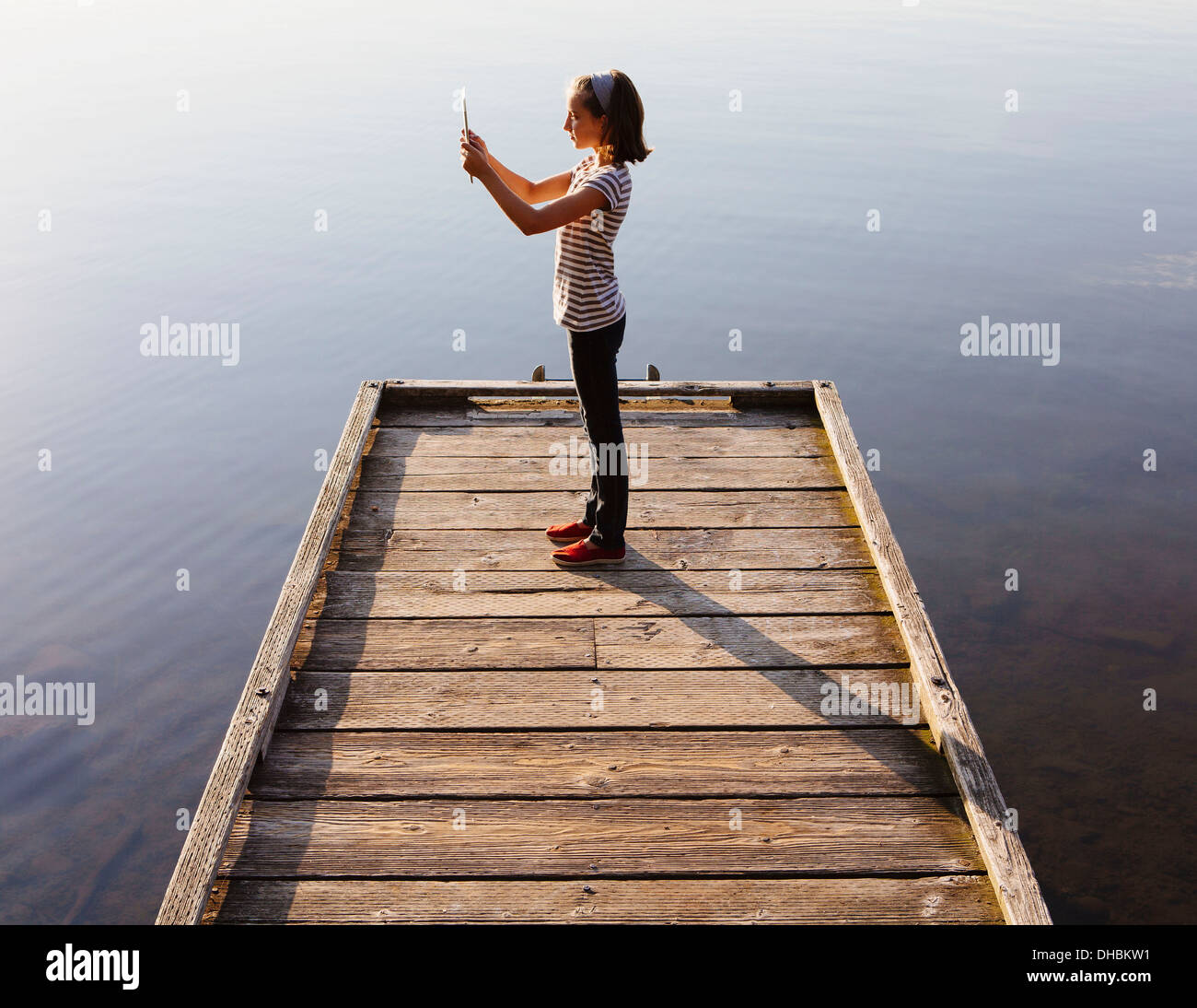 Una giovane ragazza con una tavoletta digitale di fronte a lei, in piedi su un dock in legno sopra l'acqua. Foto Stock