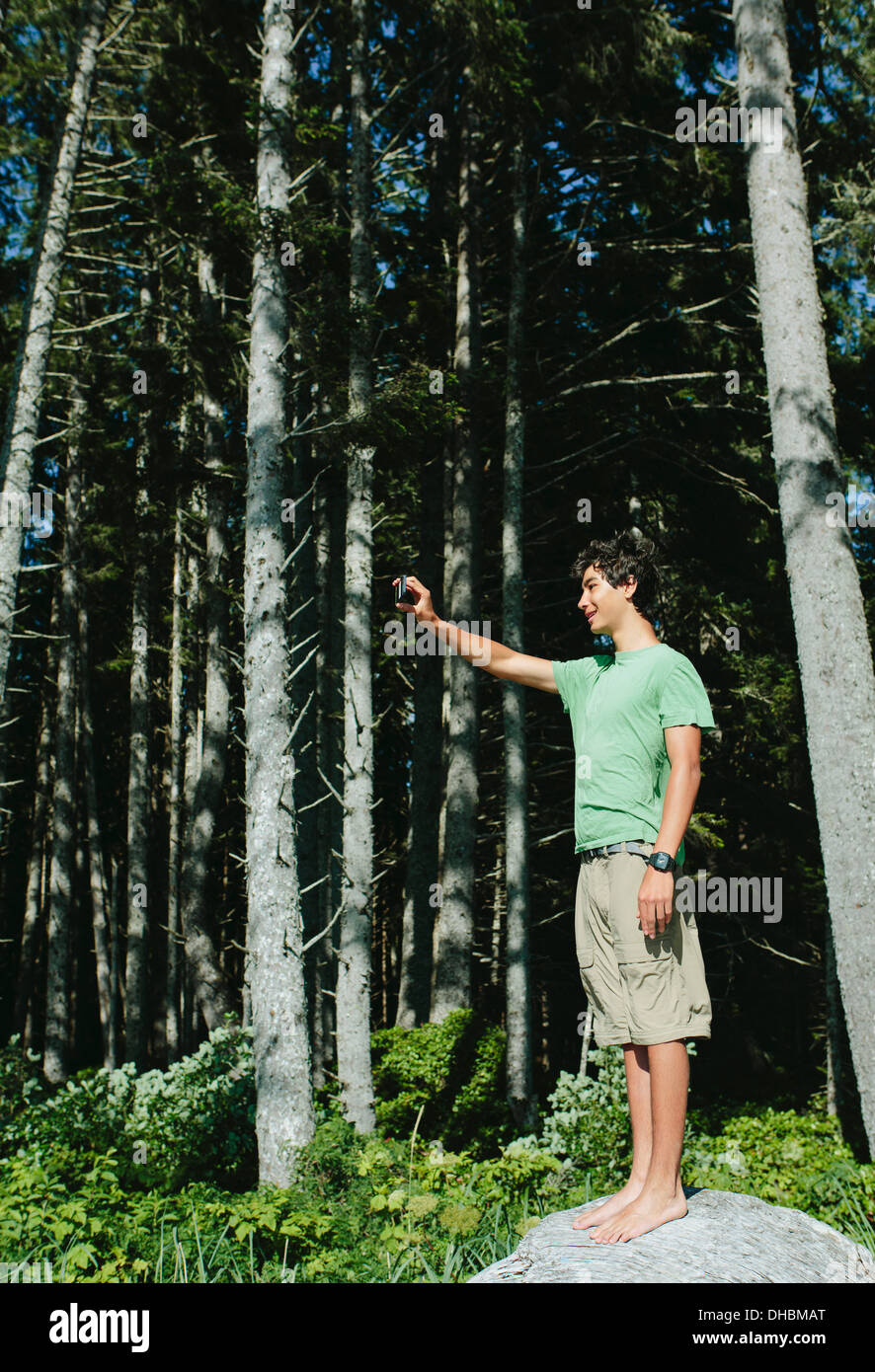 Un giovane ragazzo che sta nella foresta in possesso di una smart phone fino a prendere un selfy o una fotografia, nel parco nazionale di Olympic. Foto Stock