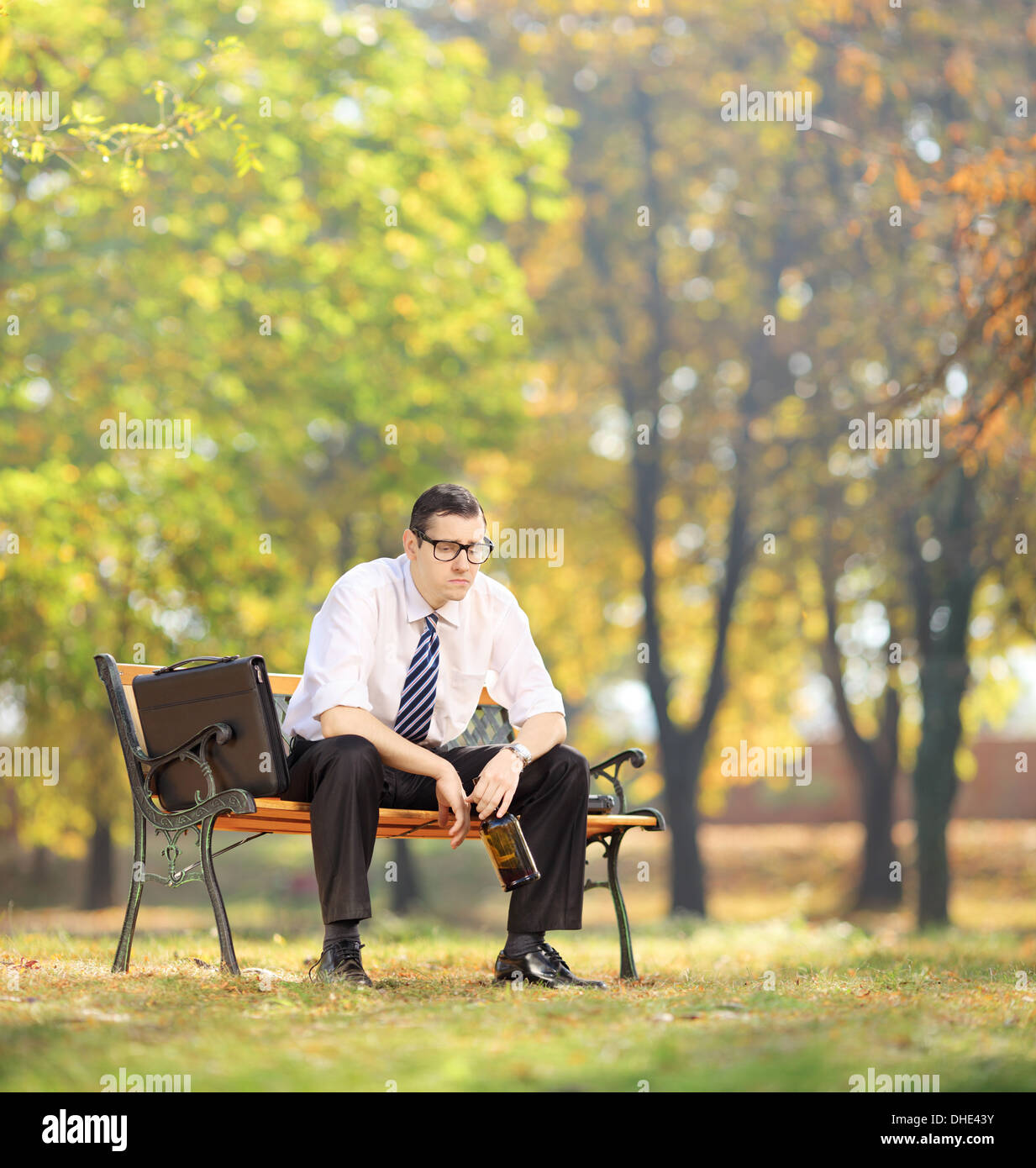 Deluso giovane imprenditore seduto su una panca in legno con bottiglia in mano in posizione di parcheggio Foto Stock