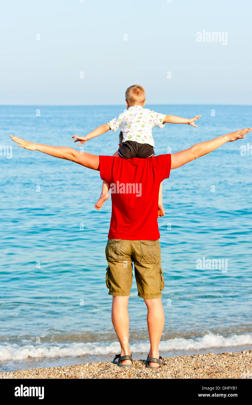 Figlio siede su di suo padre a spalle le braccia di lato contro il mare Foto Stock
