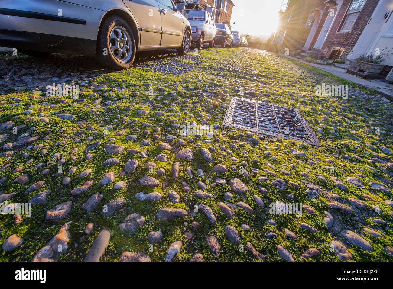 Strada di ciottoli con erba crescente tra le pietre e le macchine parcheggiate su un lato nel pomeriggio la luce diretta Foto Stock