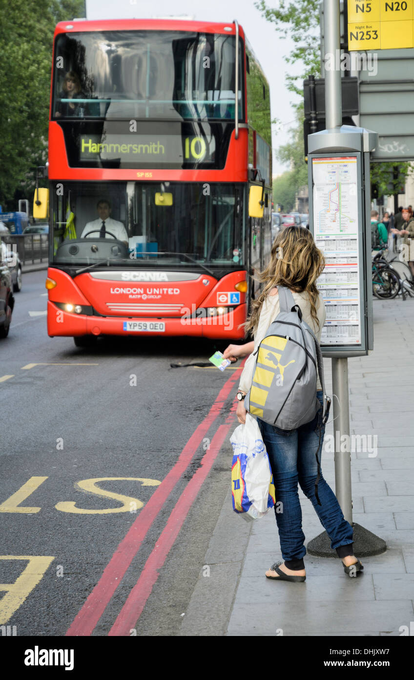 In attesa del bus: giovani femmine passeggero in un London bus stop tiene fuori il suo biglietto o abbonamento al segnale per il bus stop; donna " commuter "; bus rosso Foto Stock