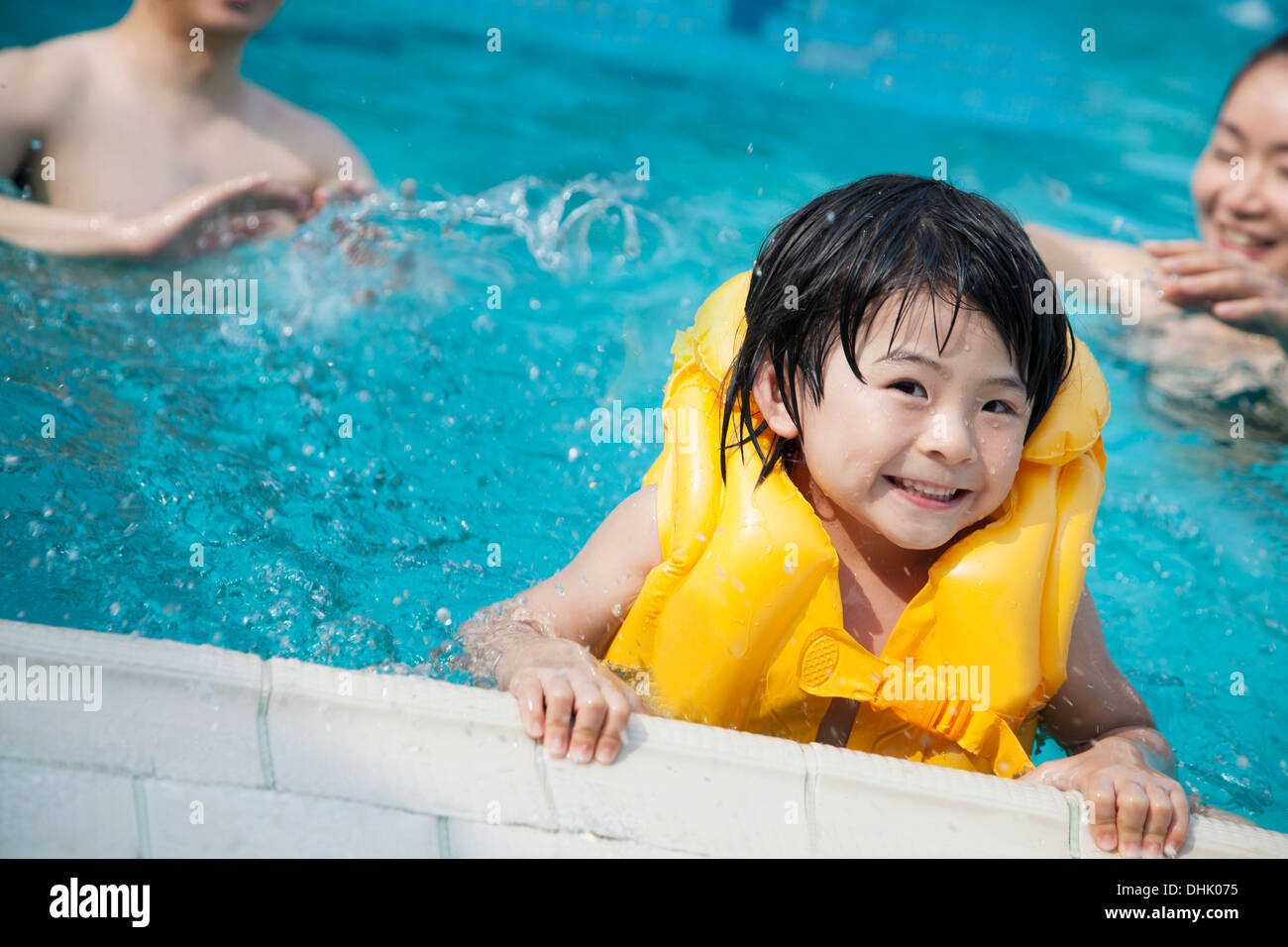Ritratto del figlio sorridente in acqua e tenendo premuto sul bordo piscine con la famiglia in background Foto Stock