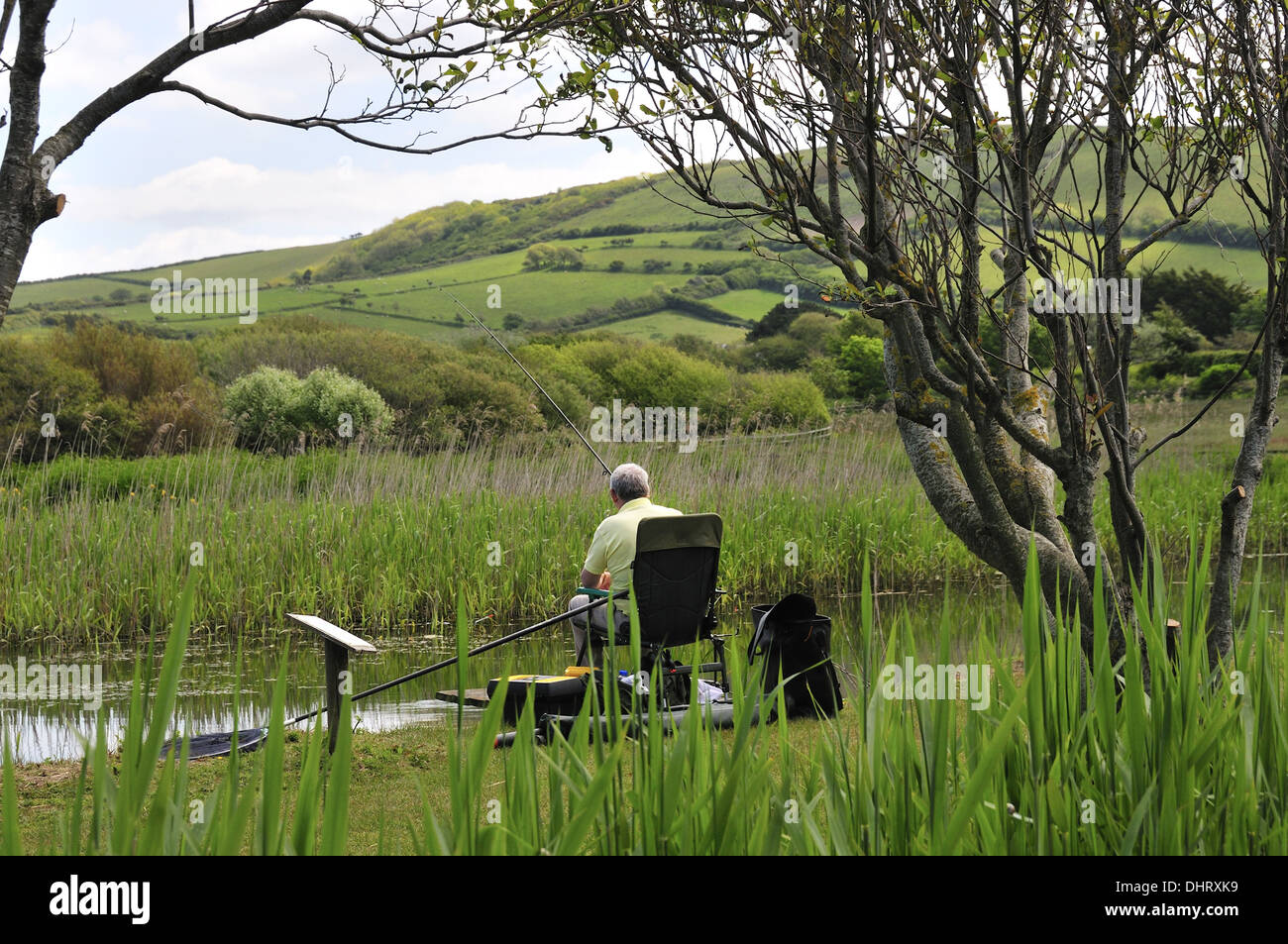 Vista posteriore dell'uomo seduto nella sedia la pesca sulle sponde di un lago del parco di Parkdean holiday park, Croyde, Devon, Inghilterra, Regno Unito Foto Stock