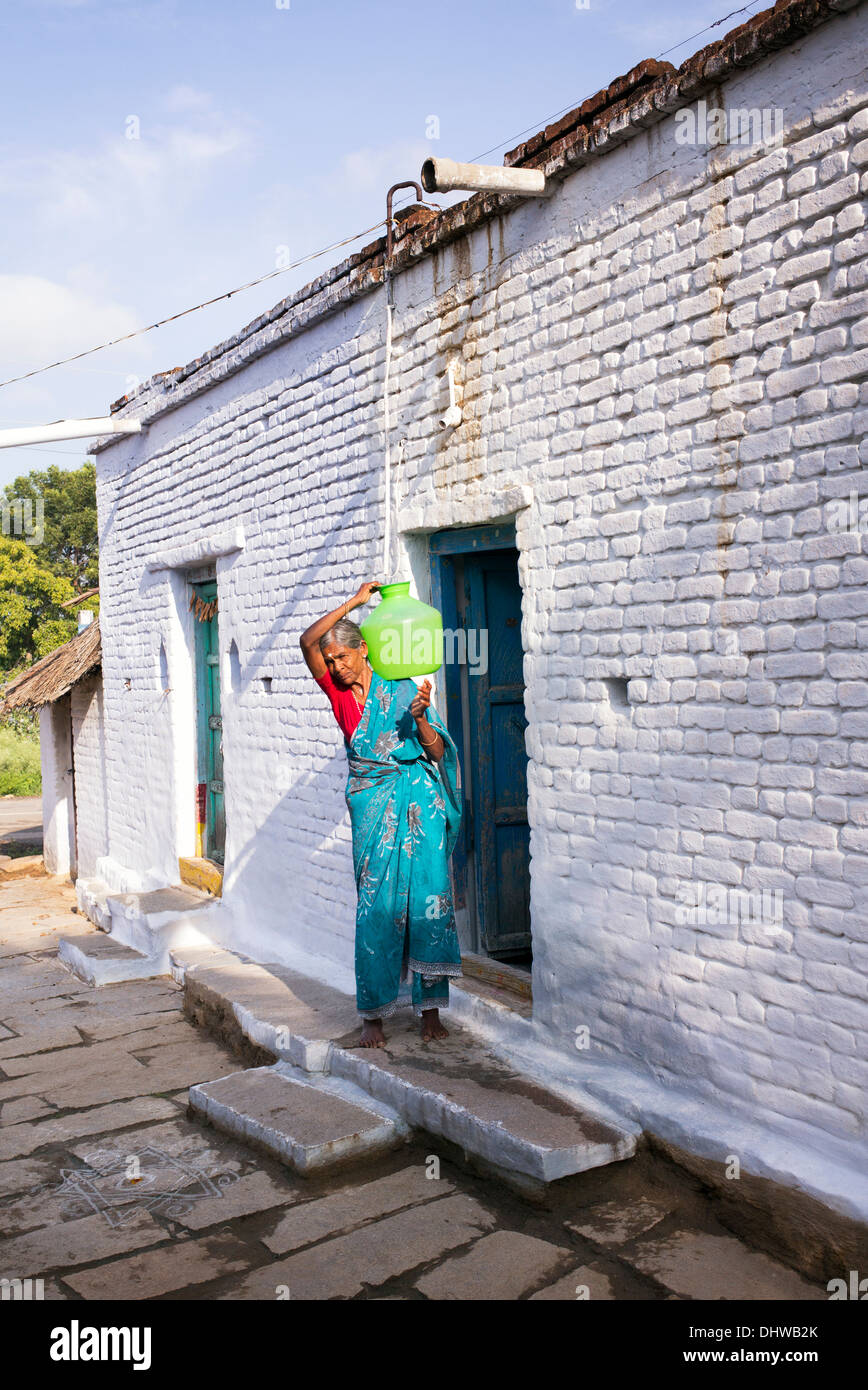 Anziani donna Indiana che porta un vaso in materia plastica con acqua da un tubo montante nel suo villaggio rurale casa. Andhra Pradesh, India Foto Stock