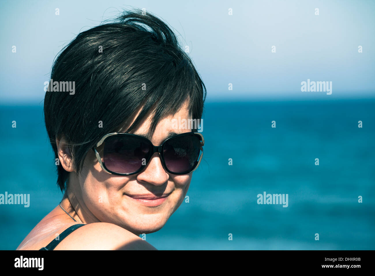 Ritratto di giovane donna sorridente in occhiali da sole rilassante sulla spiaggia e il blu del mare e del cielo. Foto Stock