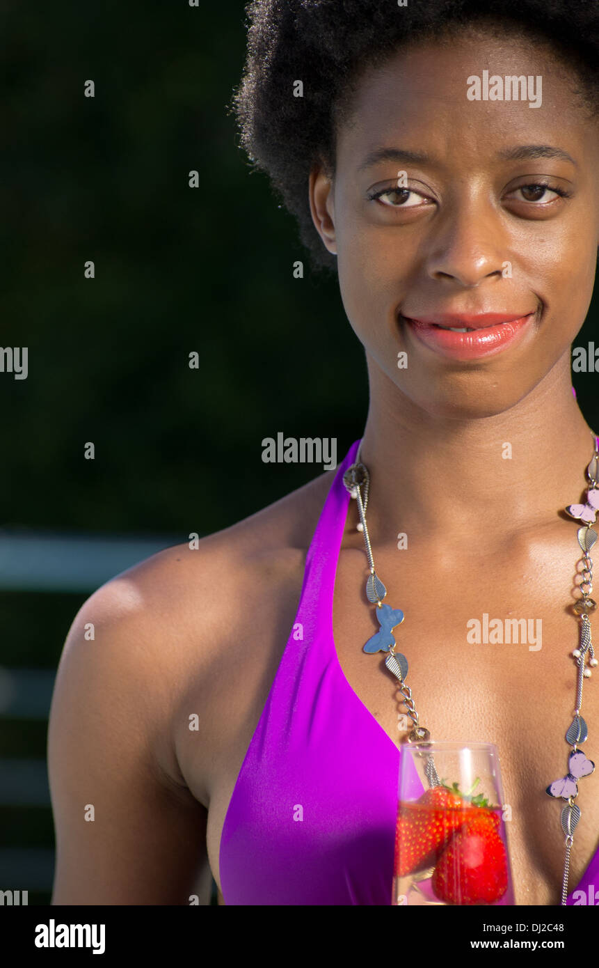 Ritratto (3/4) di un giovane africano donna americana in un viola costume da bagno con un bicchiere di champagne e fragole. Foto Stock