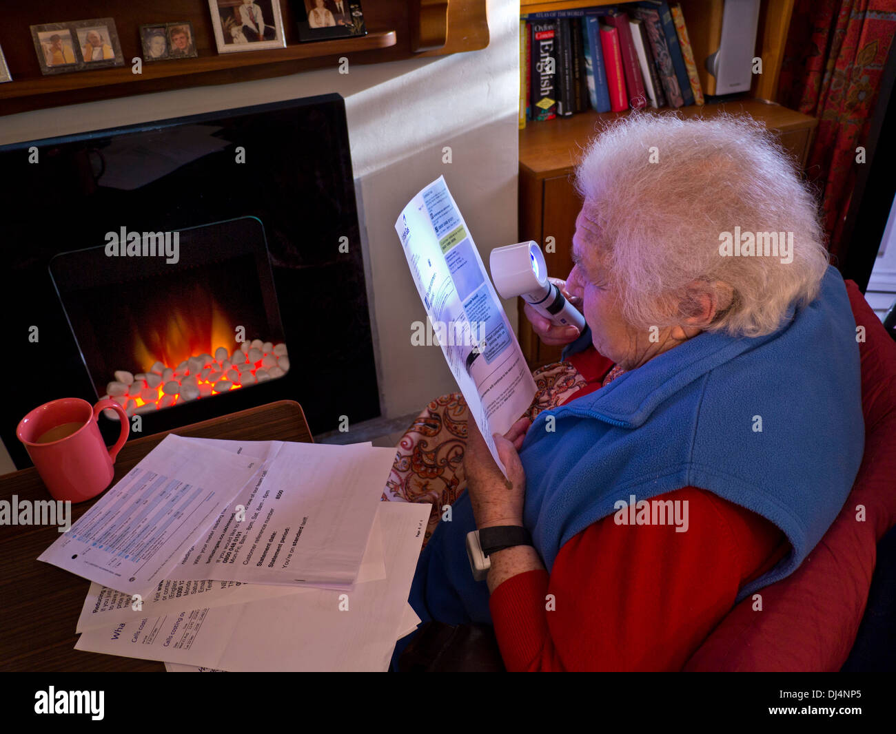 Assegno di riscaldamento anziana donna indipendente di 90 anni a casa di fronte al fuoco usando una lente di ingrandimento per leggere le sue ultime bollette energetiche domestiche Foto Stock