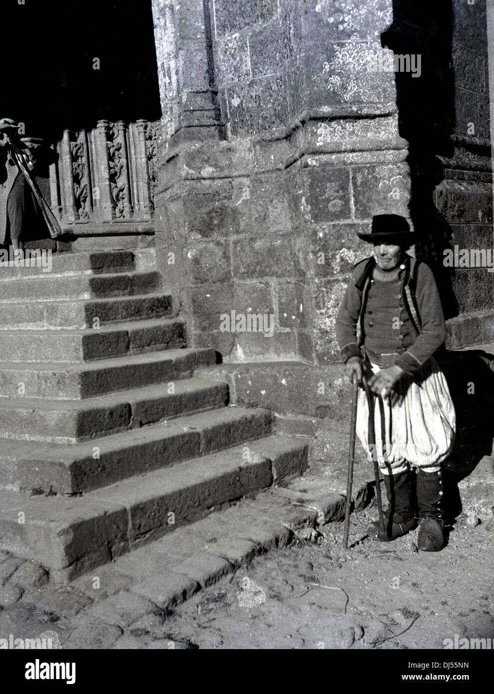 Foto storica del 1930s che mostra un anziano uomo locale vestito in costume tradizionale di pantaloni lunghi al ginocchio e tunica, in piedi con un bastone per scalini di pietra di una chiesa di Liegi, Belgio. Foto Stock