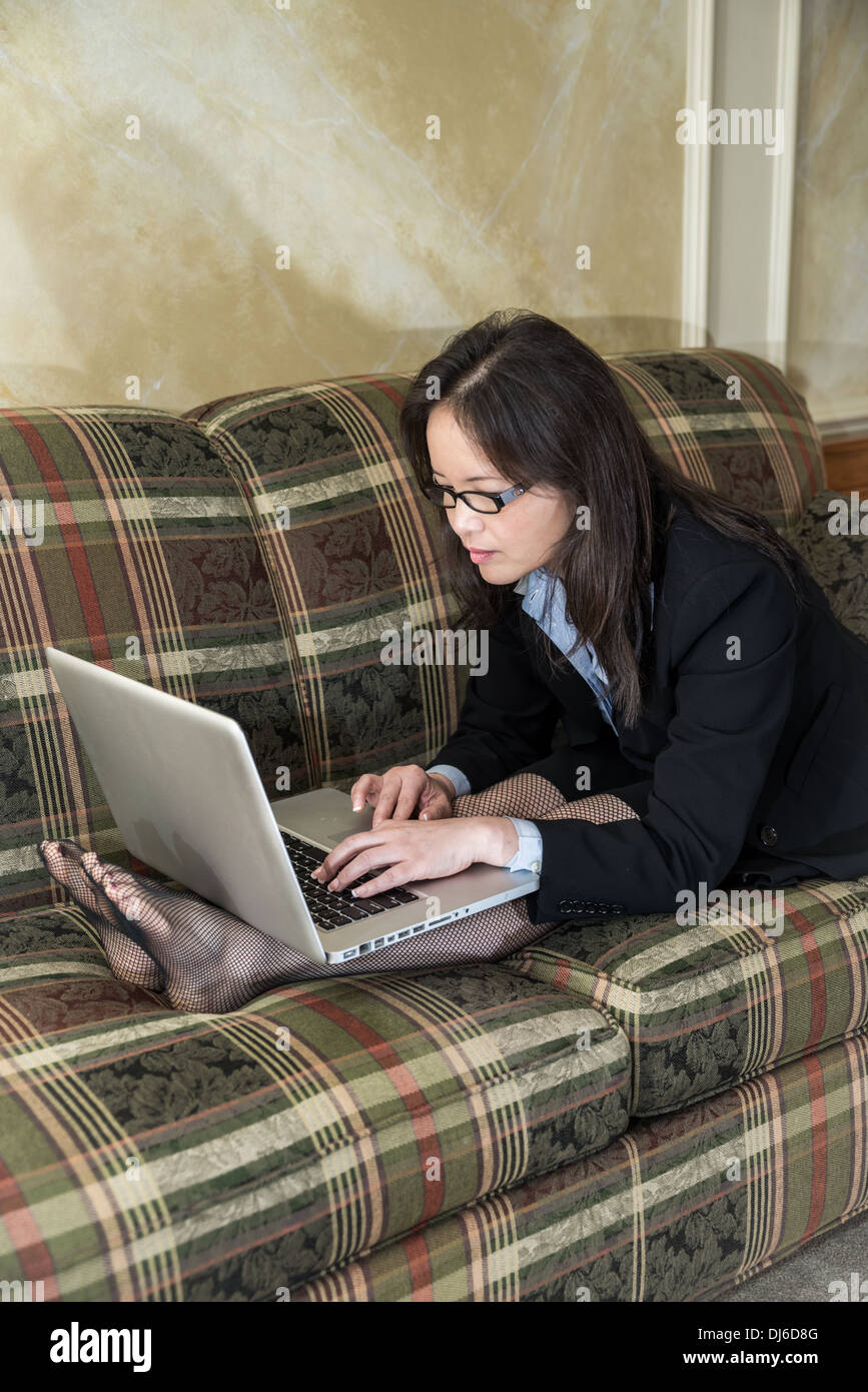 La donna nel business suit rilassante sul divano e la digitazione sul portatile a casa Foto Stock