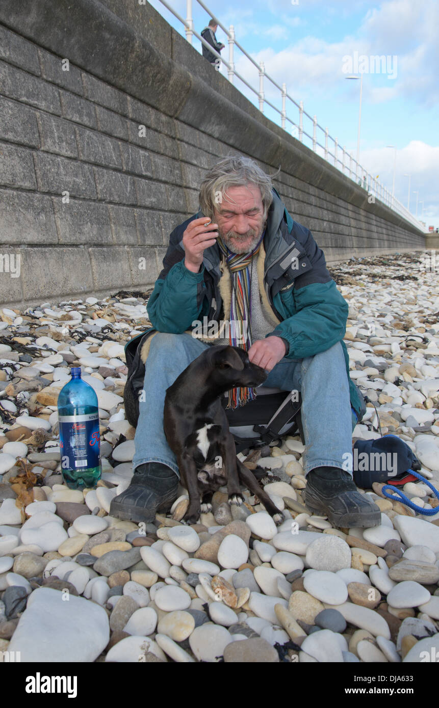 Un senzatetto uomo seduto sulla spiaggia di ciottoli con il suo cane fumare e bere Foto Stock