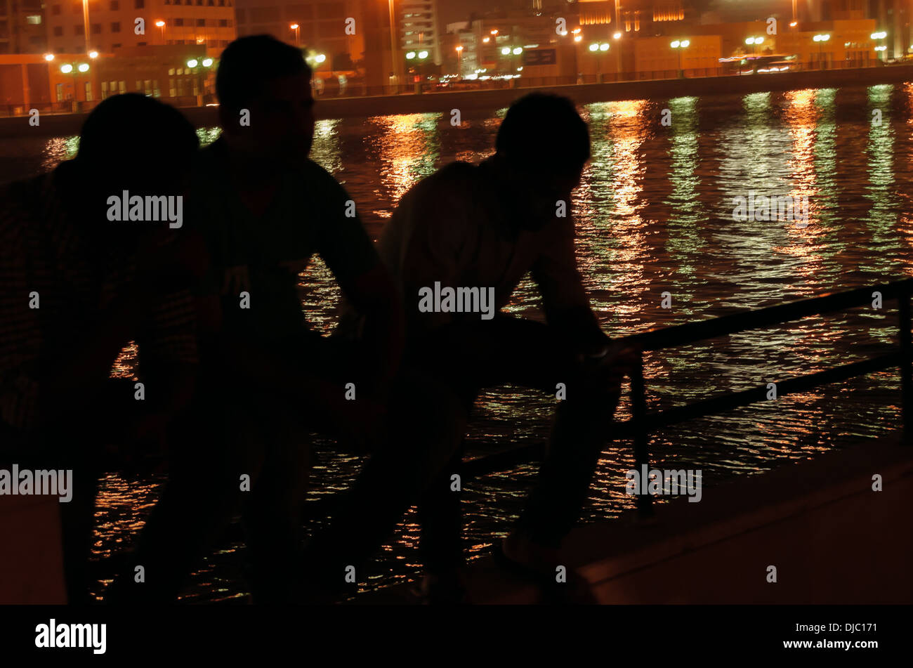 Tre uomini seduti in Dubai Creek si stagliano contro l'acqua che luccica con luci colorate di edifici vicini. Foto Stock