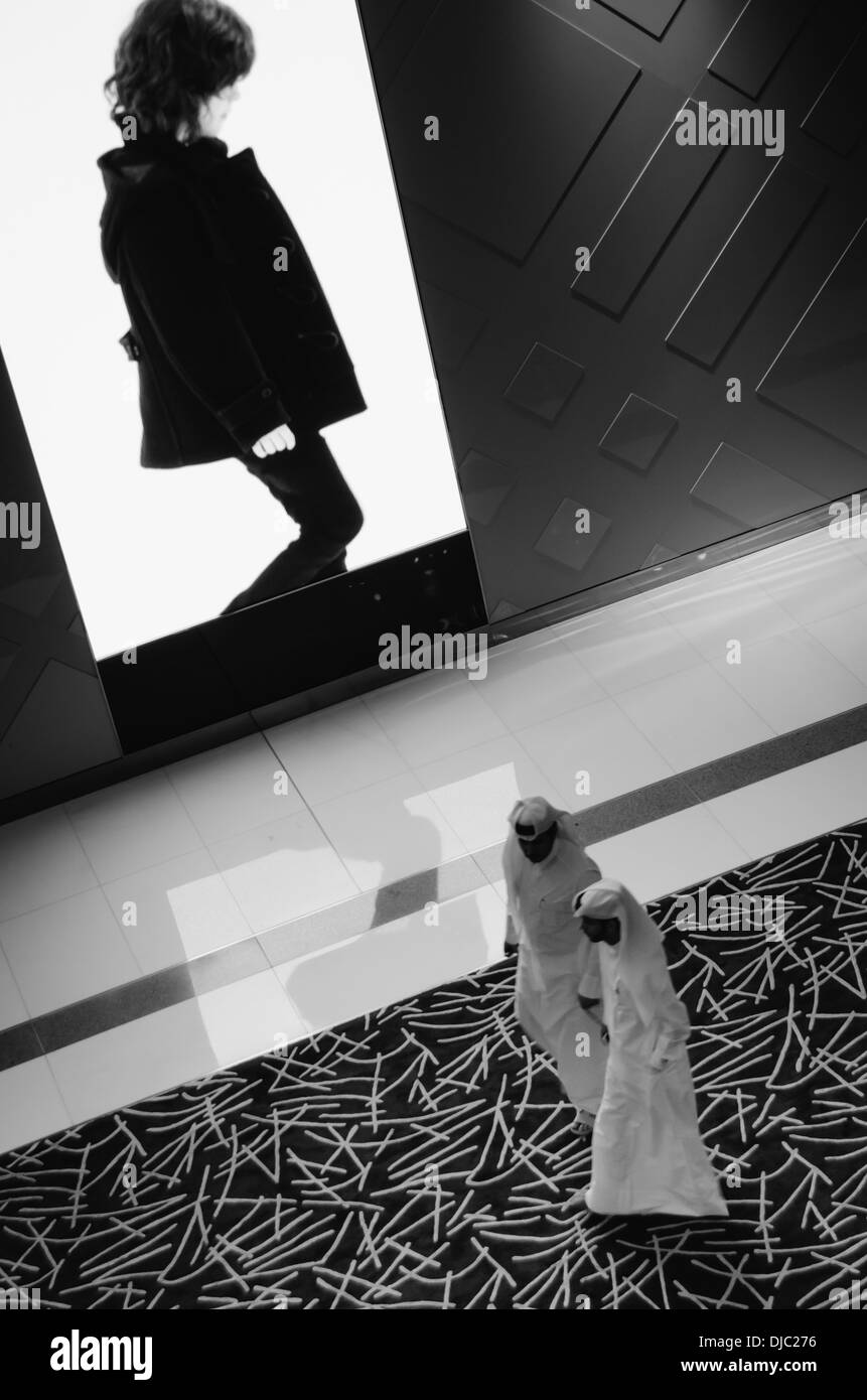 Due maschi arabi nel Dubai Mall a piedi nella parte anteriore di un grande schermo a LED con un modello occidentale a camminare in direzione opposta. Foto Stock