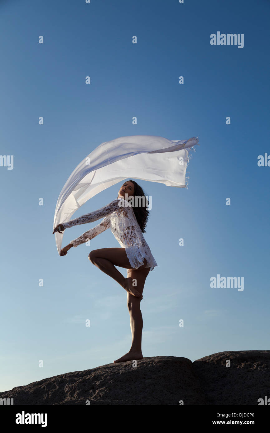 Modello femminile sventolare una lunga sciarpa bianca in aria contro un profondo cielo blu mentre si fermò a piedi nudi su una roccia che indossa bikini bianco Foto Stock