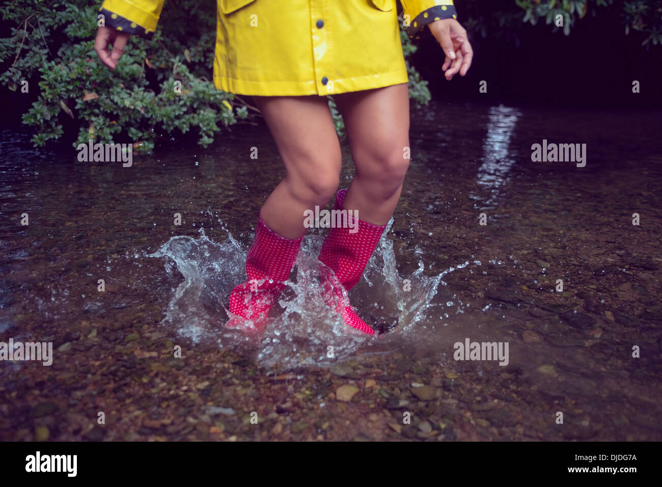 Donna in impermeabile giallo e rosso gumboots saltando in acqua Foto Stock