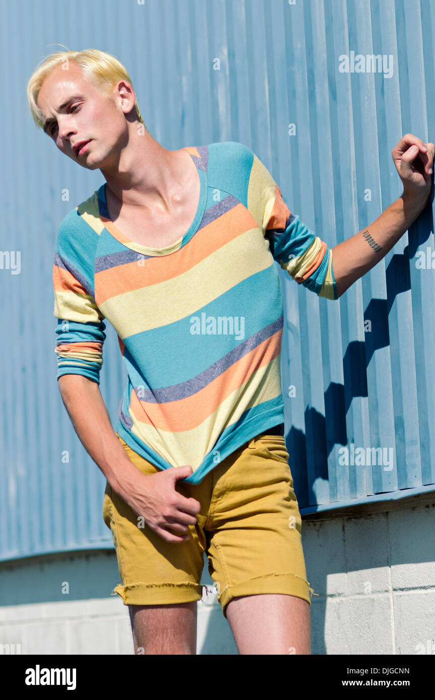Un giovane uomo, bionda, che pongono, indossa una camicia striscia e pantaloncini, pendente, mano sulla parete metallica. Un estate fashion concept. Foto Stock